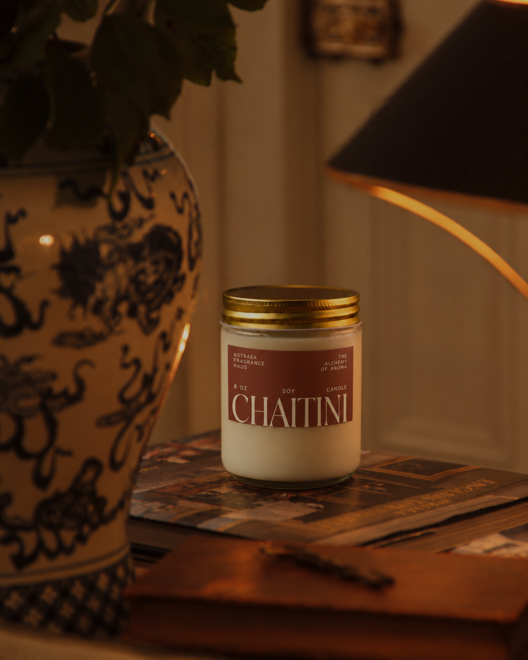 A white soy candle in a glass jar with a gold lid, labeled "Chaitini" by Astraea Fragrance Haus, placed on a wooden surface near a black lampshade.