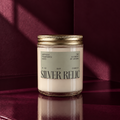 Silver Relic soy candle in a clear glass jar with a gold lid, label with black text, set against a dark red background.